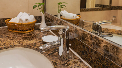 A fragment of a white washbasin. Shiny stainless steel faucet.  There is a soap dish on the countertop, towels, toiletries in a wicker basket, a green plant in a vase. Reflection in the wall mirror.