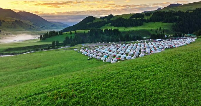 Aerial shot of a large traditional yurt campsite situated in lush green mountain valley at sunrise in Xinjiang, China.