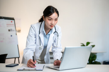 Asian female doctor holding pen and standing leaning over table to look at laptop in diagnosis room.