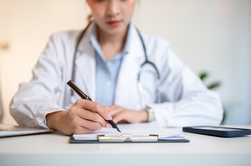 Asian female doctor holding pen writing in paper on clipboard with laptop on table in diagnosis room