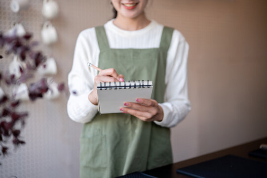 Asian female barista holding pencil writing in notebook taking customer's order at cafe counter bar.