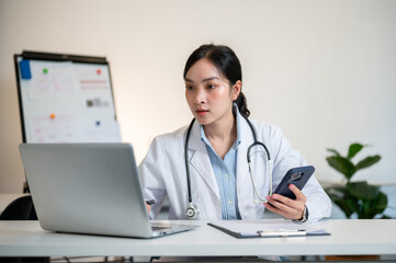 Asian female doctor holding smartphone and looking at laptop on working table in diagnosis room.