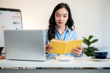 Female office worker or businesswoman reading yellow cover book with laptop aside on working table.