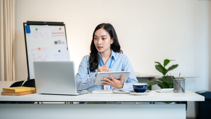 Female office worker businesswoman holding stylus writing in tablet while looking at laptop on table
