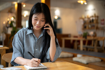 Asian woman talking on phone holding pen writing in notebook with calculator on wooden table in cafe
