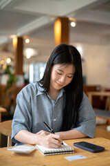 Asian woman smiling while holding pen writing in notebook with calculator and phone on cafe's table.
