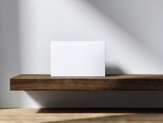A blank white card rests on a dark brown wooden shelf against a minimalist white wall, sunlit