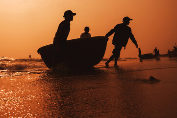 Silhouettes of Vietnamese fishermen with traditional round basket boat on beach in a fishing...
