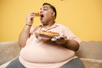 Hungary overweight fat indian man eating cheese pizza. Plus size guy enjoy fatty junk food. Obese male taking a bite of high calorie meal. Obesity and weight gain.