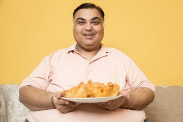 Happy overweight indian man holding plate of samosa snack looking at camera. Plus size guy enjoy fried junk food.