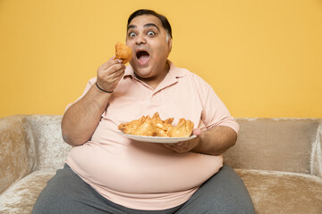 Hungary overweight fat indian man eating unhealthy samosa junk food. Plus size guy eating fried snack while sitting on couch at home.. Obesity and weight gain. Obese male craving high calorie meal.