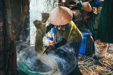 Vietnamese woman worker in straw hat dyes reeds in boiling water on a stove at a woven mat factory...