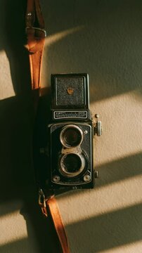 Vintage twin lens reflex camera with brown leather strap, rests on a beige surface. Sunlight casts dramatic parallel shadows. Dark, moody tones. 