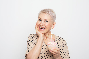 Happy senior female celebrating birthday with cupcake with candle over light grey background. Party and people concept.