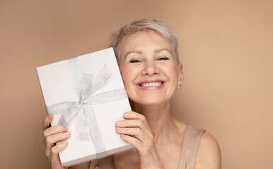 Portrait of cheerful positive glad charming aged woman having gift box over beige backgrou