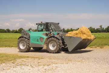 Obraz premium Front loader with a bucket full of hay on a gravel road in a sunny rural field, concept of agriculture, farming industry, heavy machinery