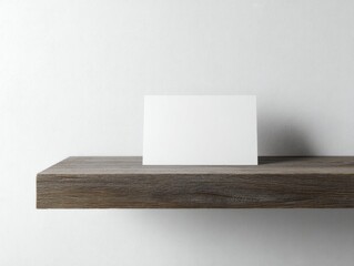 Blank white card rests on a rustic, dark-grey wood shelf against a minimalist white wall