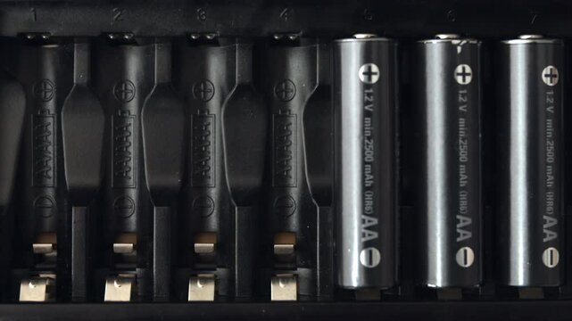 Placing stick-shaped AAA batteries into a black electronic device with a male hand from overhead view. Clean minimal background emphasizes focused insertion moment.
