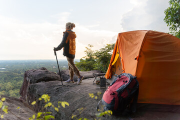 A woman is standing on a rock with a backpack and a tent. She is wearing an orange jacket