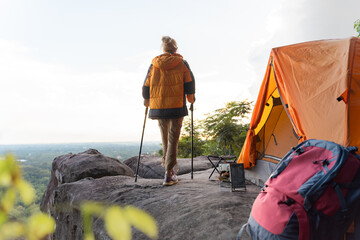 A woman is walking on a rocky mountain with a backpack and a tent. She is wearing a yellow jacket