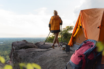 A woman is standing on a rock with a backpack and a tent. She is wearing a yellow jacket