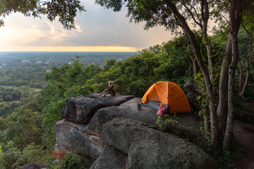 A person is sitting in front of a tent. The tent is orange. The person is wearing a yellow shirt