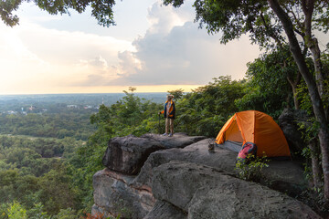 A man is standing on a rock with a tent and a backpack. The tent is orange and the backpack is red