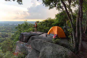 A man is standing on a rocky hillside with his backpack and orange tent set up. The tent is pitched...