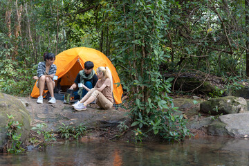 Three people are sitting around a tent in the woods. One of them is reading a book. The tent is orange