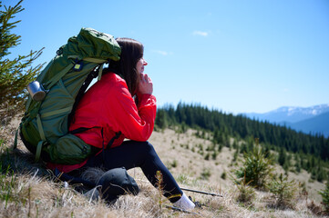 Female hiker resting on grassy mountain slope with trekking poles. Woman in red jacket enjoying...