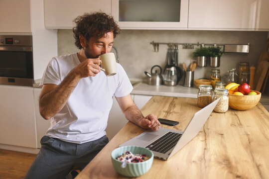 A man sits at a wooden kitchen counter, sipping coffee from a mug, focused on his laptop. A bowl of cereal and fresh fruit are nearby, creating a cozy breakfast atmosphere.