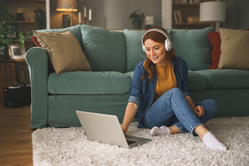 Woman with long red hair enjoys time at home, seated on soft rug with laptop in front. She wears...