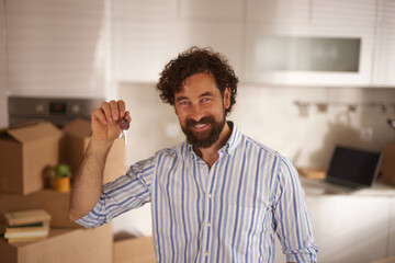 A cheerful man stands in his new home, holding keys and smiling widely with packed boxes around...