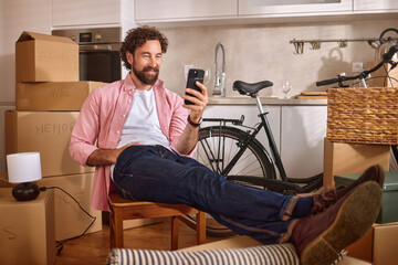 Relaxing in a new home, a man enjoys his phone while sitting on a chair with his feet up. Nearby...