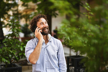 A man with a beard smiles while talking on his phone, standing among lush plants in an urban space,...
