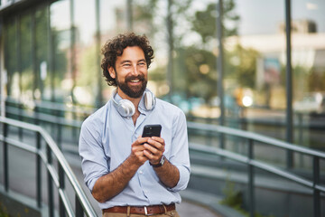 A man with curly hair and headphones stands outside a modern building, smiling while engaged with...