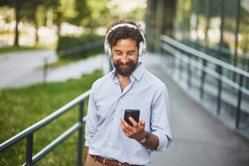 Man dressed in a light shirt listens to music with headphones while using his smartphone, walking...