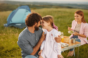 A family of three is having a cheerful picnic in a green field. Parents are interacting warmly with...
