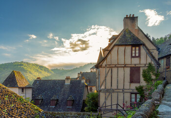 Panoramic view of the historic village of Conques with rustic medieval houses with the mountains in the background illuminated by the golden light of sunset under a sky with backlit clouds.