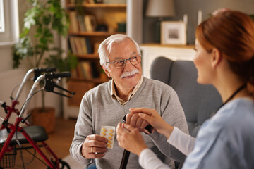 An elderly man holding a package of pills receives care at home from a woman in medical scrubs. She gently holds his hand in a comforting gesture in his living room.