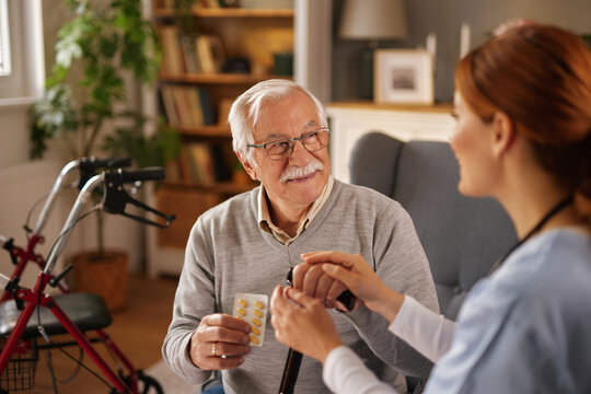 An elderly man holding a package of pills receives care at home from a woman in medical scrubs. She gently holds his hand in a comforting gesture in his living room.