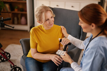 An elderly woman, seated in a chair, receives medication from a home nurse, who also provides...