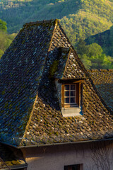 Traditional dormer window on a rustic mossy slate roof in the French mountain village of Conques, France, with golden sunset sunlight illuminating the wooden window.