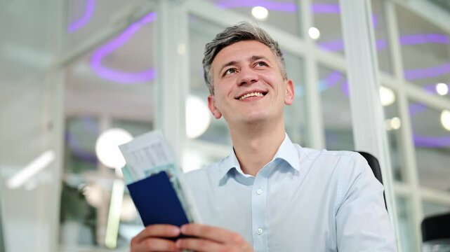 Cheerful man, dressed in light blue shirt, holds passport and travel documents in bright office. Professional smiles happily, looking forward to exciting journeys.