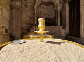 Photo of a brass candle holder in sand-filled tray at the entrance of Panagia Ekatontapiliani church in Parikia, Paros Island, framed by Byzantine arches and historic stonework.