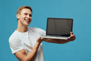 smiling young man hold laptop presenting blank screen wearing white t-shirt isolated blue...