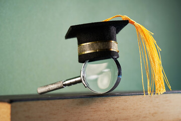 small mortarboard and magnifying glass on top of hard cover book against chalkboard