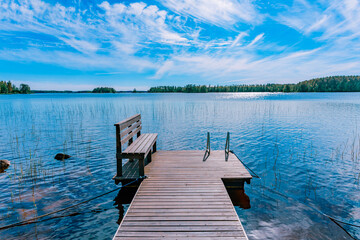 Obraz premium Wooden pier on blue lake with a bench and a ladder in Finland. The water is calm and the sky is blue