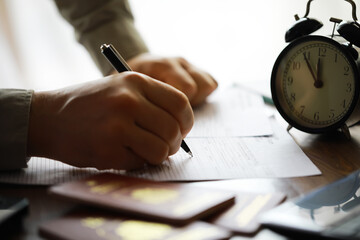 Close-Up of Hand Writing on Paper with Passports and Alarm Clock on Desk - Deadline Concept