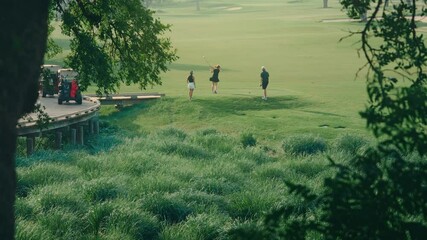 Distant shot through trees capturing one male and two female golfers on the tee box preparing to tee off. Scenic golf course setting with natural framing, bright weather, and a calm outdoor atmosphere